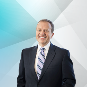 Joel Susco, smiling in a professional headshot. He is wearing a black suit with a white collar shirt and a stripe blue tie. The background consist of different shades of blue.