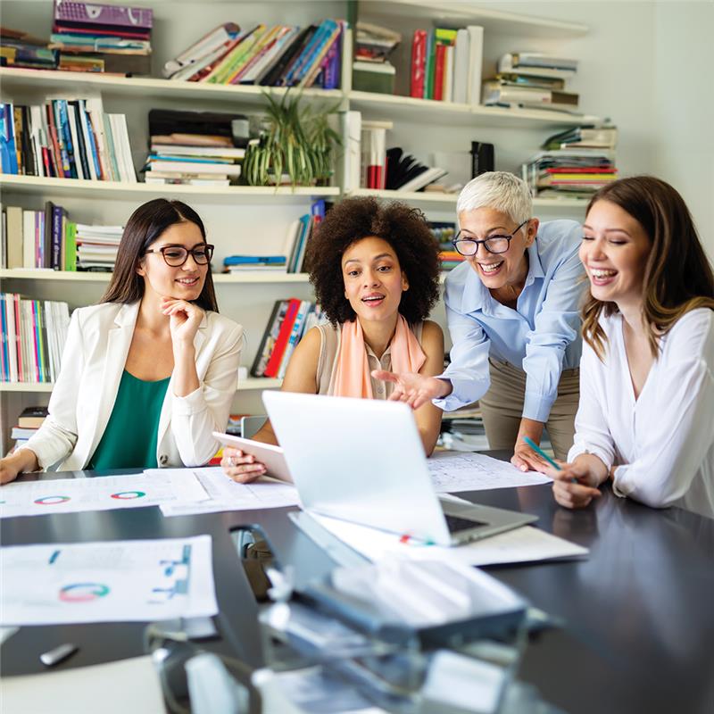 Four women engaged in discussion around a table, with a laptop open in front of them, fostering collaboration and ideas.