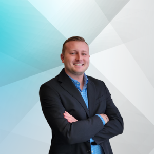 Ryan Kelly is smiling in a professional headshot. The background is a blue color. He is wearing a black blazer with a blue collar long sleeve shirt.